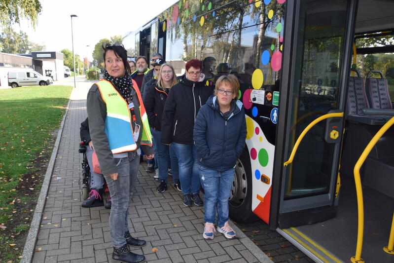 Eine Frau mit einer Warnweste steht mit einer Gruppe Menschen an einem Bus.
